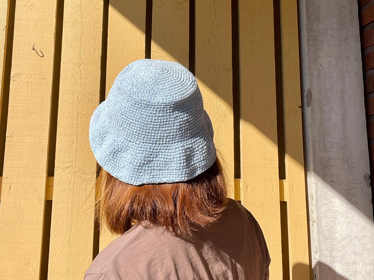 En person med hår till axlarna som står med ryggen mot kameran, personen bär en virkad bucket hat i ljusblå färg.