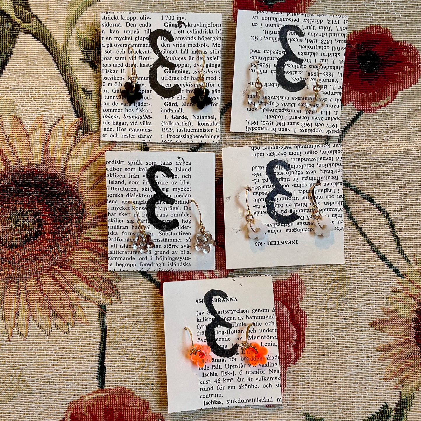 Fem par örhängen i guldpläterat metall, alla med hängen som ser ut som små blommor i olika färger. Svarta blommor, genomskinliga blommor, blommor i spegelglas, vita blommor och orange blommor.