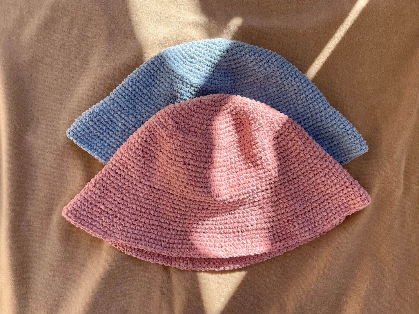 En bild på en blå bucket hat och en rosa bucket hat som ligger på en beige filt i solen. 