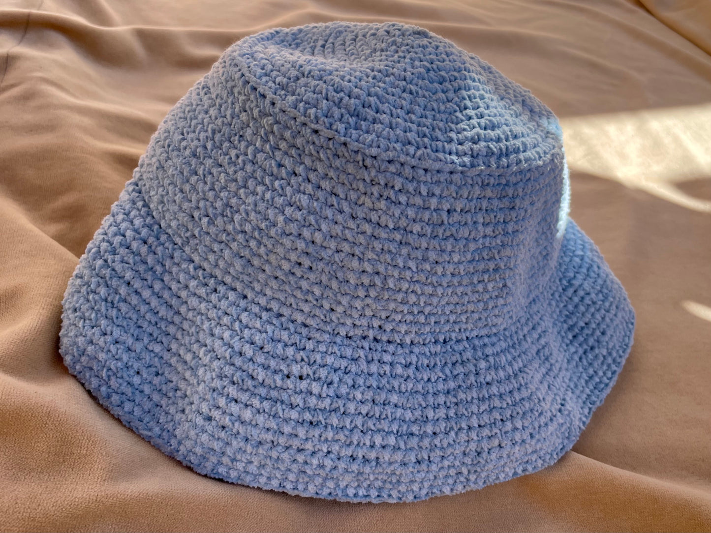 En närbild på en virkad ljusblå bucket hat som ligger på en beige filt i solen. 